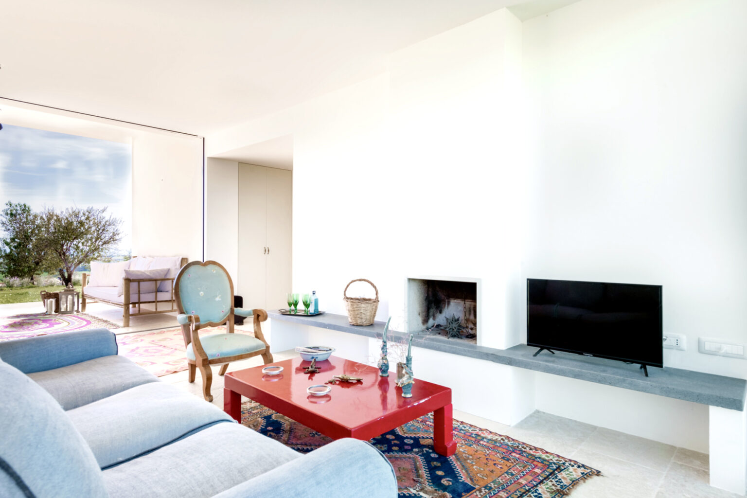 Modern living room featuring a light blue sofa, red coffee table, and a flat-screen TV. Large windows offer natural light and a view of the outdoors. The space is decorated with a colorful rug and a stylish armchair, creating a cozy atmosphere. | Only on SopranoVillas