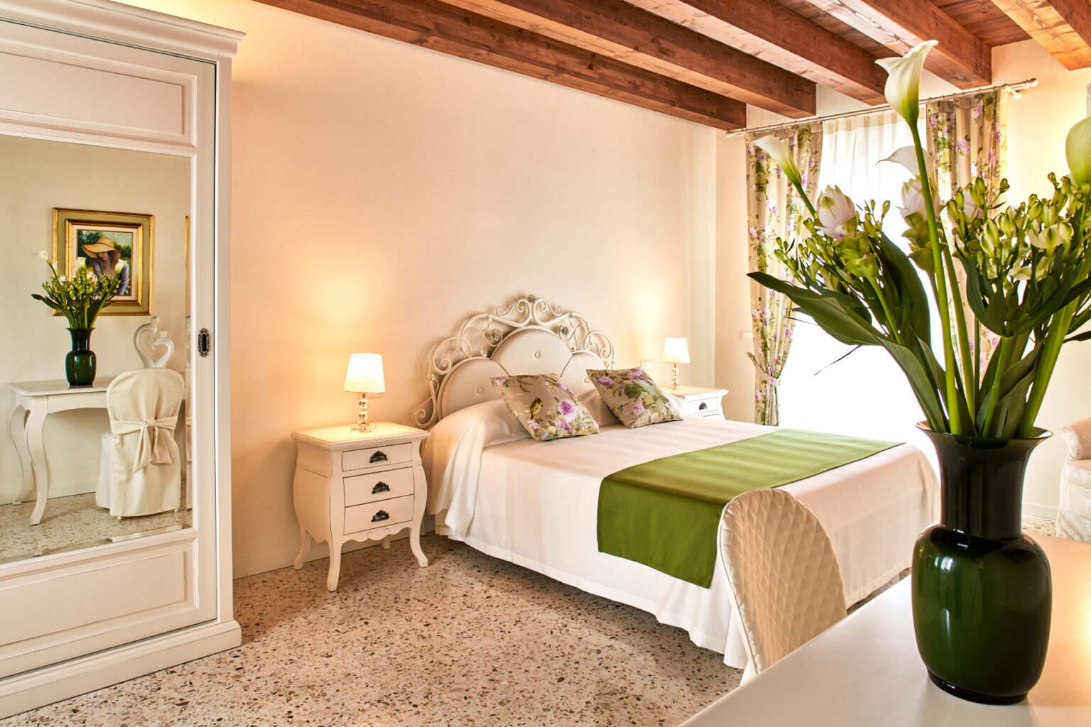 Cozy and elegantly designed bedroom featuring a double bed with green accents, a vintage white dresser, and a large mirror. Natural light floods the room, highlighting the floral decor and wooden beams on the ceiling. Ideal for a relaxing retreat. | Only on SopranoVillas