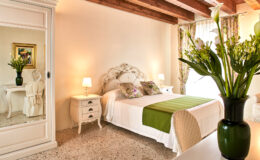 Cozy and elegantly designed bedroom featuring a double bed with green accents, a vintage white dresser, and a large mirror. Natural light floods the room, highlighting the floral decor and wooden beams on the ceiling. Ideal for a relaxing retreat.