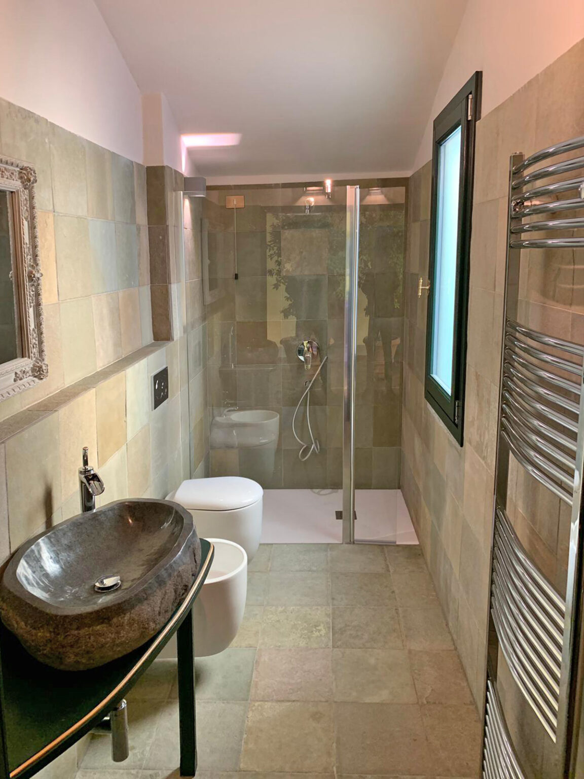Modern bathroom featuring a glass shower enclosure, stylish round sink, sleek toilet, and a heated towel rail, with natural stone walls and neutral color palette. | Only on SopranoVillas