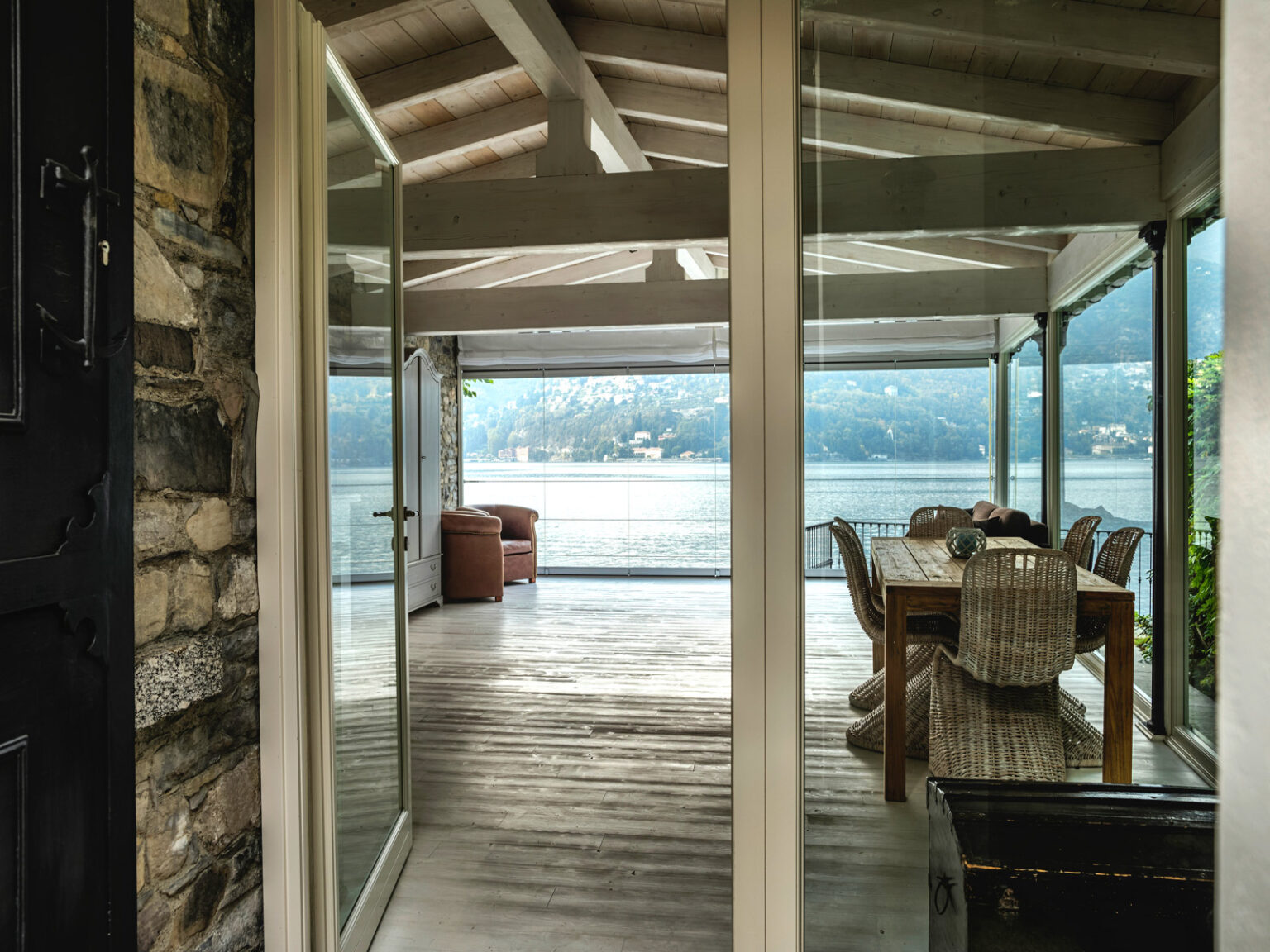 Modern outdoor dining area with a wooden table and wicker chairs, featuring a scenic view of the lake through glass doors, surrounded by a covered patio. | Only on SopranoVillas
