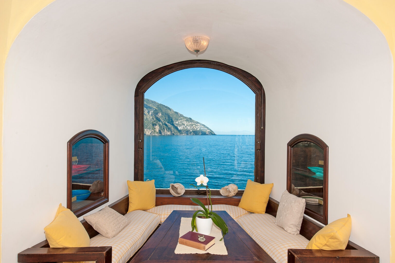 Cozy living space with a panoramic ocean view, featuring a comfortable seating area with yellow cushions, a wooden table, and a potted orchid, framed by an arched window that overlooks the serene coastline. | Only on SopranoVillas