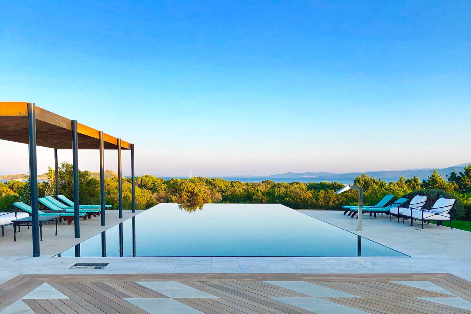Infinity pool overlooking lush greenery and a clear blue sky, with lounge chairs and a shaded seating area in a serene outdoor setting. | Only on SopranoVillas