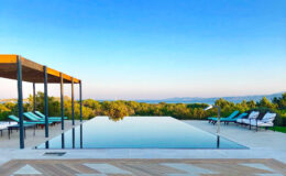 Infinity pool overlooking lush greenery and a clear blue sky, with lounge chairs and a shaded seating area in a serene outdoor setting.