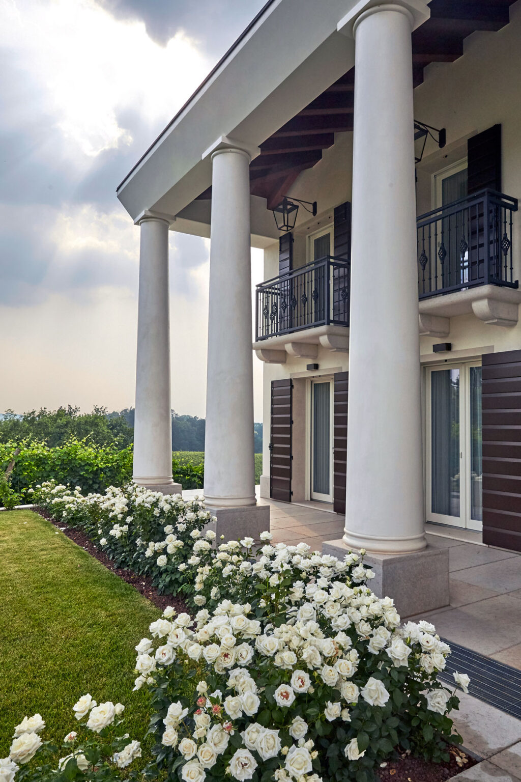 Elegant modern home with white columns and a balcony, surrounded by lush greenery and blooming white roses, showcasing a serene outdoor setting. | Only on SopranoVillas