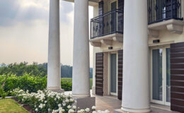 Elegant modern home with white columns and a balcony, surrounded by lush greenery and blooming white roses, showcasing a serene outdoor setting.
