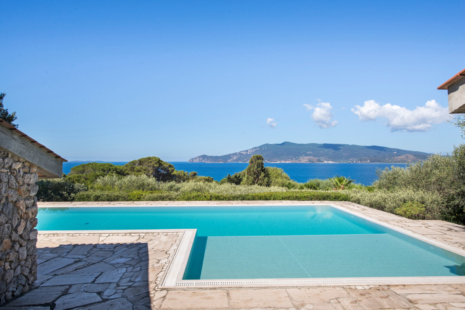Modern villa with a stunning infinity pool overlooking the ocean and island in the distance, surrounded by lush greenery and clear blue skies. | Only on SopranoVillas