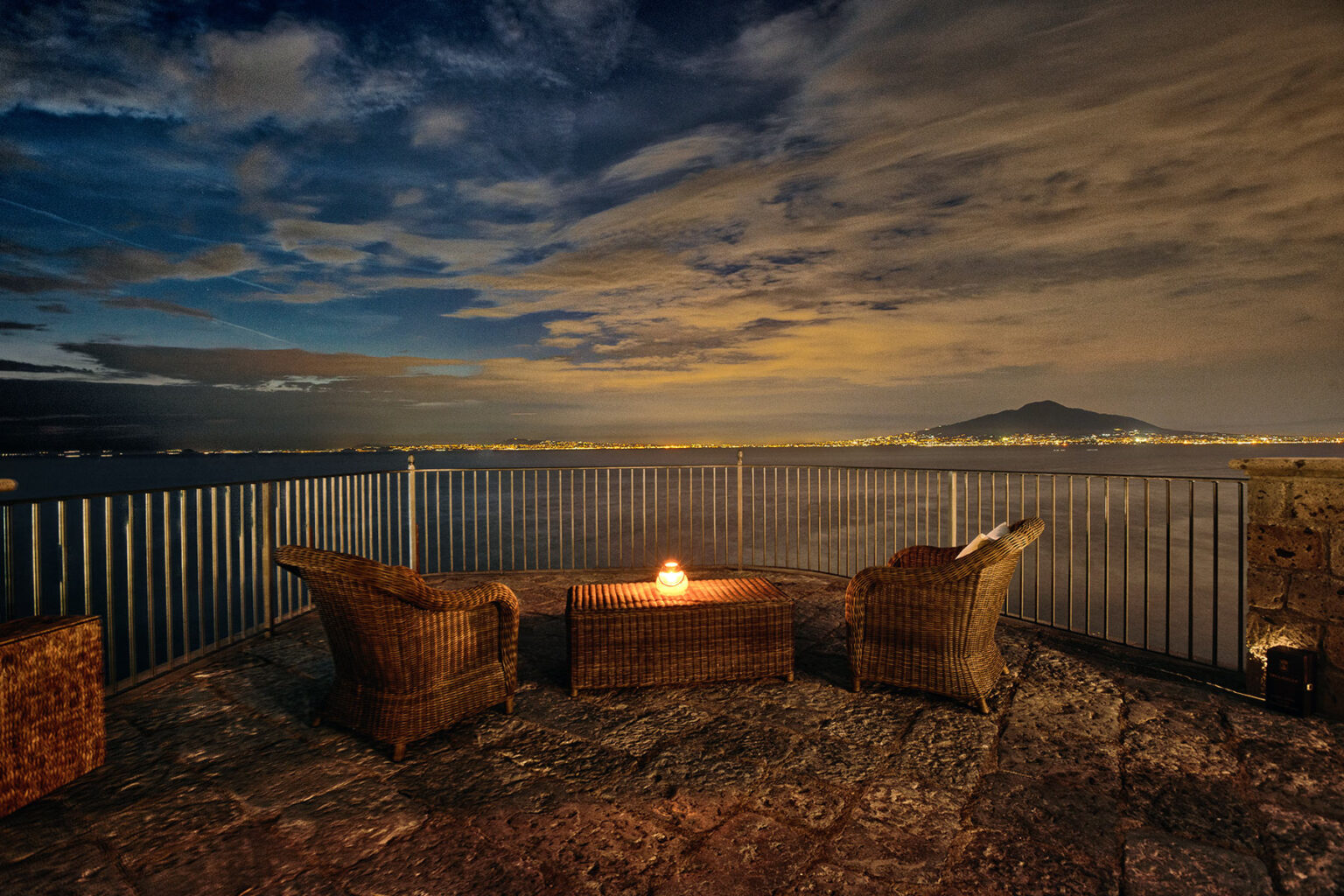 Cozy outdoor seating area with two wicker chairs and a small fire pit overlooking a tranquil body of water at night, with a scenic view of distant mountains and a dramatic sky filled with clouds. | Only on SopranoVillas