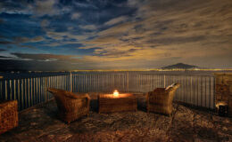 Cozy outdoor seating area with two wicker chairs and a small fire pit overlooking a tranquil body of water at night, with a scenic view of distant mountains and a dramatic sky filled with clouds.