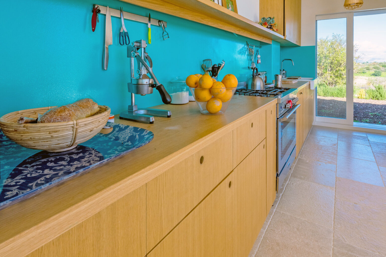 Modern kitchen with turquoise walls, wooden cabinetry, and a countertop featuring a bowl of oranges and a basket of bread. Natural light floods in from a nearby sliding door, enhancing the inviting atmosphere. | Only on SopranoVillas