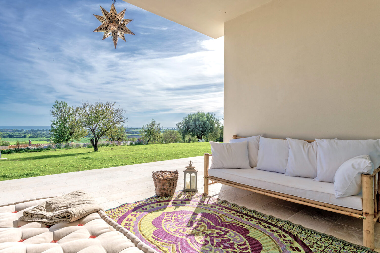 Cozy outdoor seating area with a white sofa and decorative pillows, featuring a colorful rug and a lantern, overlooking a lush green landscape under a bright blue sky. Ideal for relaxation and enjoying nature. | Only on SopranoVillas