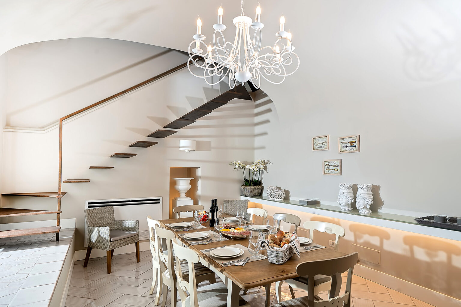 Modern dining area featuring a wooden table set for a meal, elegant chandelier, and stylish staircase. The bright space includes decorative elements and a cozy atmosphere, ideal for gatherings. | Only on SopranoVillas