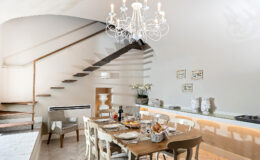 Modern dining area featuring a wooden table set for a meal, elegant chandelier, and stylish staircase. The bright space includes decorative elements and a cozy atmosphere, ideal for gatherings.