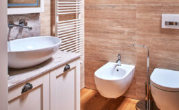 Modern bathroom interior featuring a stylish round sink, a wall-mounted bidet, and a sleek toilet. The warm wooden flooring complements the elegant beige wall tiles, creating a cozy yet sophisticated atmosphere.