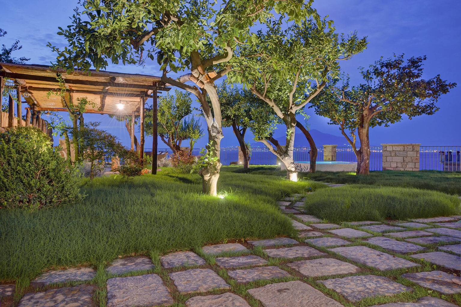Serene outdoor garden at dusk featuring lush green grass, illuminated trees, and a stone pathway leading to a wooden pergola overlooking the water. | Only on SopranoVillas