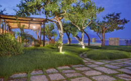 Serene outdoor garden at dusk featuring lush green grass, illuminated trees, and a stone pathway leading to a wooden pergola overlooking the water.