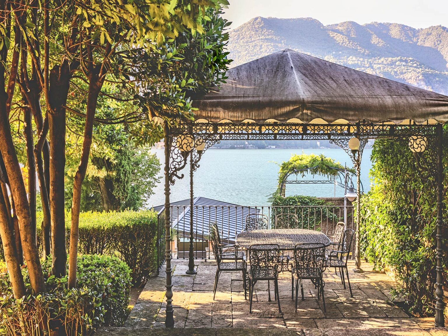 Scenic garden terrace with a gazebo overlooking a serene lake, surrounded by lush greenery and mountains in the background, perfect for outdoor dining and relaxation. | Only on SopranoVillas