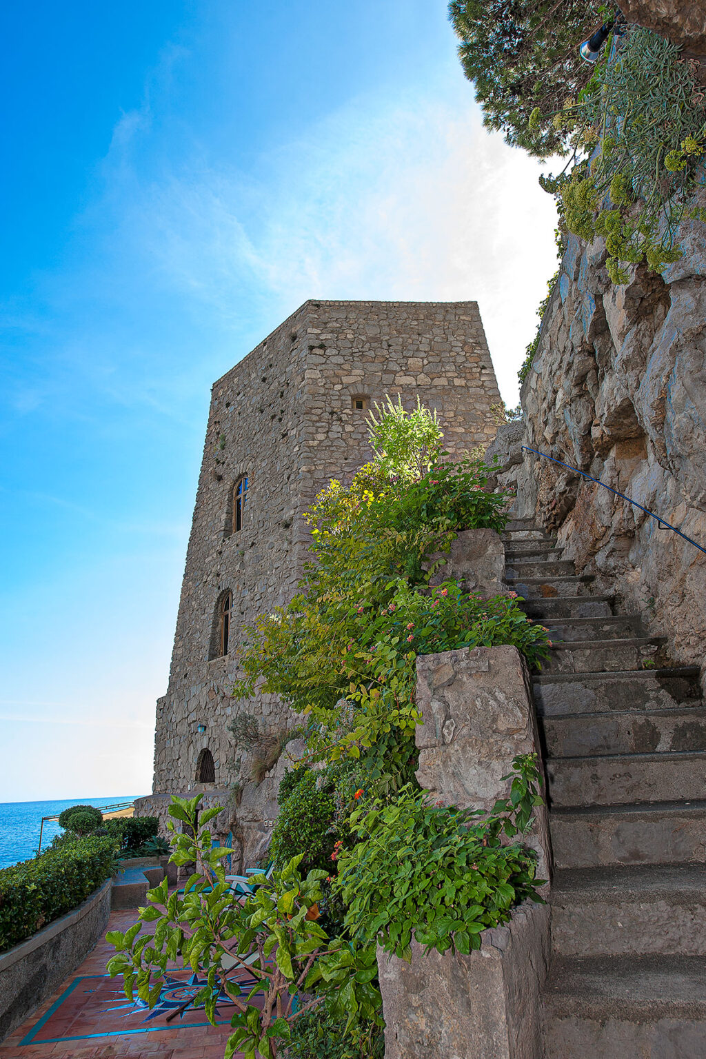 Stone tower with greenery and stone steps under a blue sky, showcasing historical architecture and natural elements. | Only on SopranoVillas