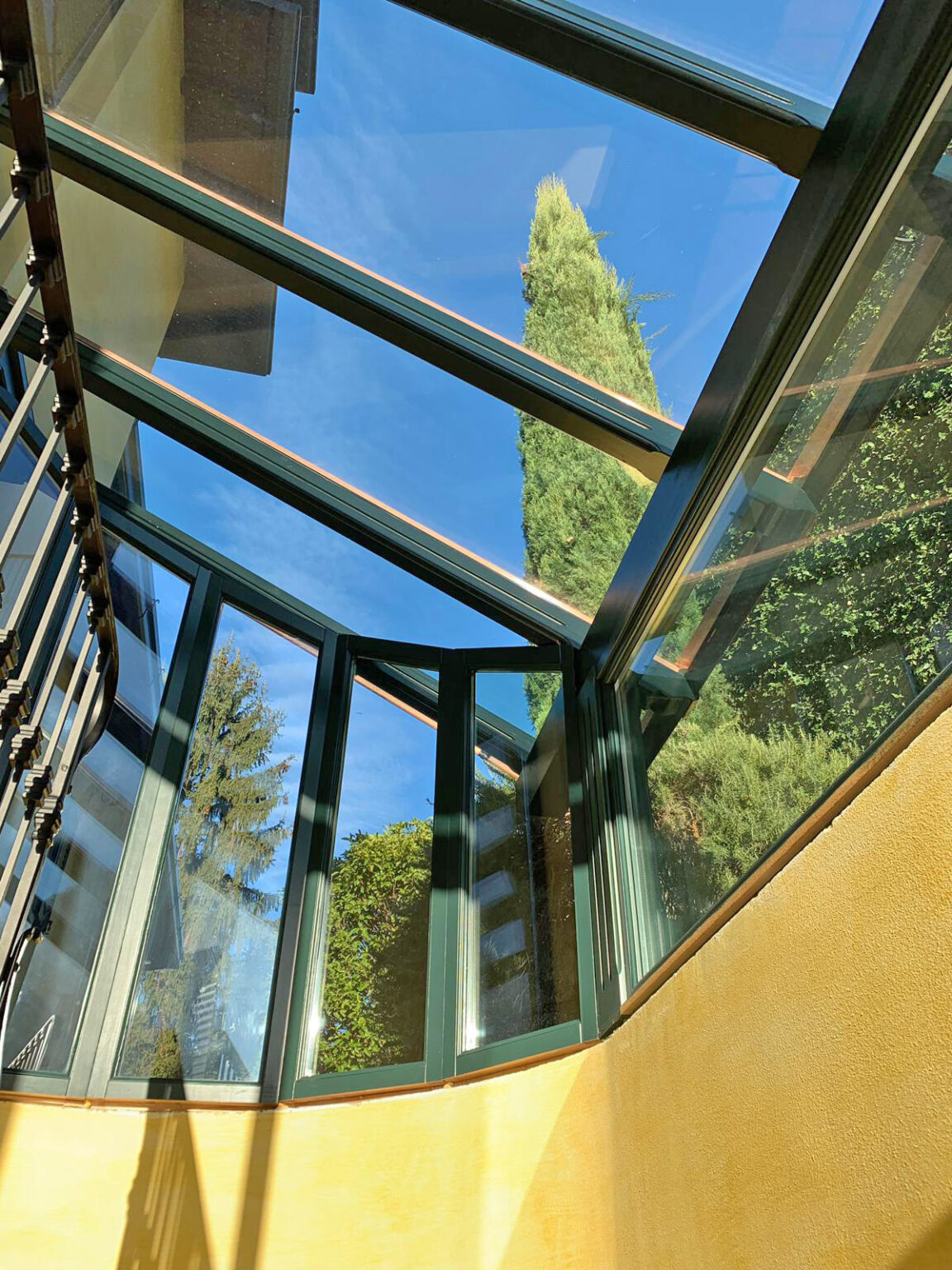 Interior view of a modern glass structure with a clear blue sky visible through the ceiling, surrounded by lush greenery. The design features angular glass panels and a contemporary aesthetic, highlighting natural light and outdoor elements. | Only on SopranoVillas