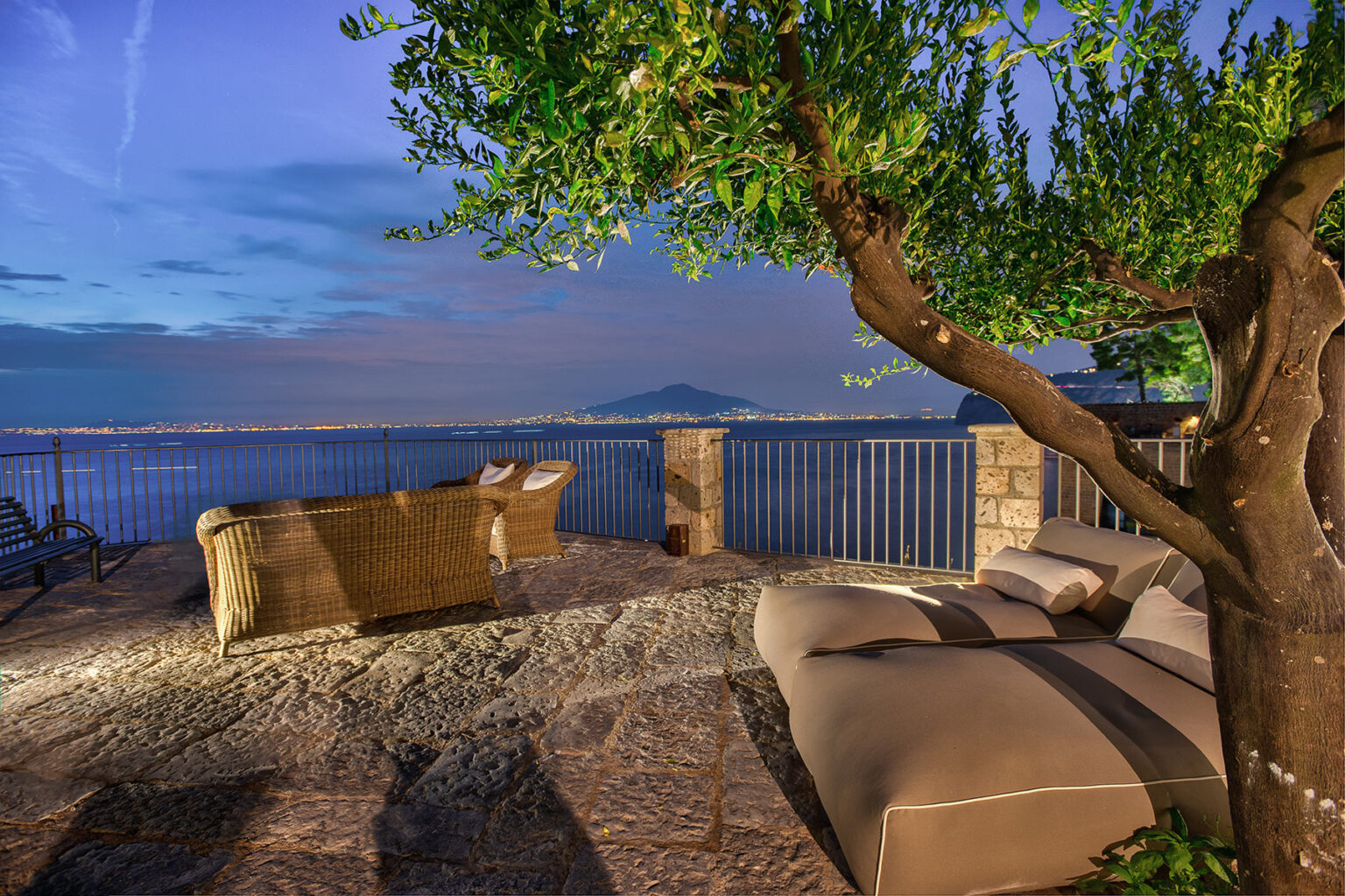 Cozy outdoor seating area with a view of the sea at dusk, featuring a comfortable lounge chair and a tree providing shade, surrounded by stone flooring and a railing. | Only on SopranoVillas