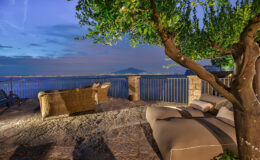 Cozy outdoor seating area with a view of the sea at dusk, featuring a comfortable lounge chair and a tree providing shade, surrounded by stone flooring and a railing.