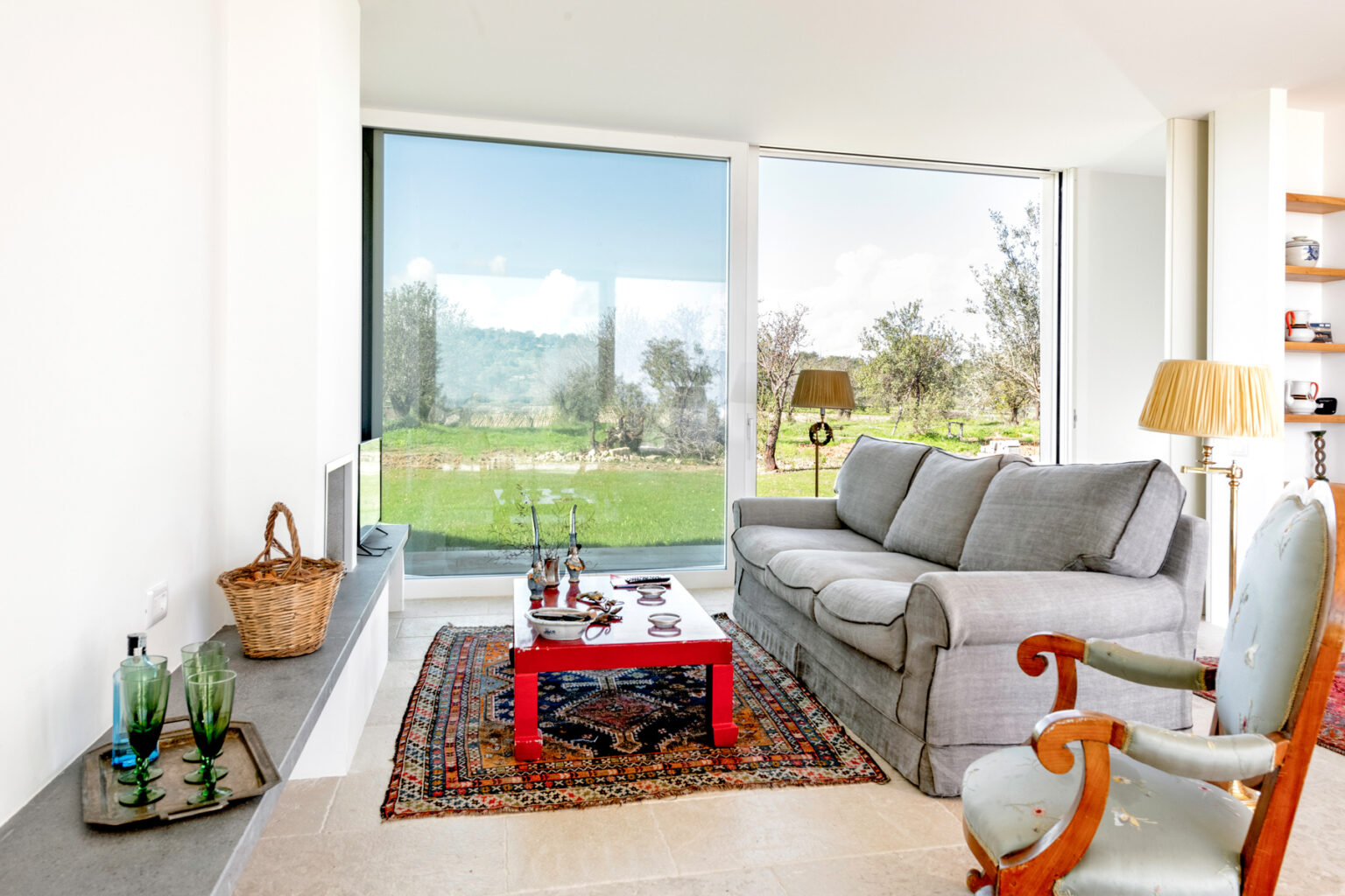 Cozy living room featuring a gray sofa, a red coffee table, and a large window with a view of greenery. The space includes decorative items like a woven basket and colorful glass vases, creating a warm and inviting atmosphere. | Only on SopranoVillas