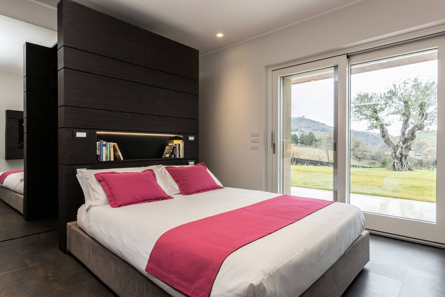 Modern bedroom featuring a queen-sized bed with pink accent pillows, stylish dark wood paneling, and large sliding glass doors opening to a scenic outdoor view. | Only on SopranoVillas