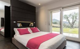 Modern bedroom featuring a queen-sized bed with pink accent pillows, stylish dark wood paneling, and large sliding glass doors opening to a scenic outdoor view.