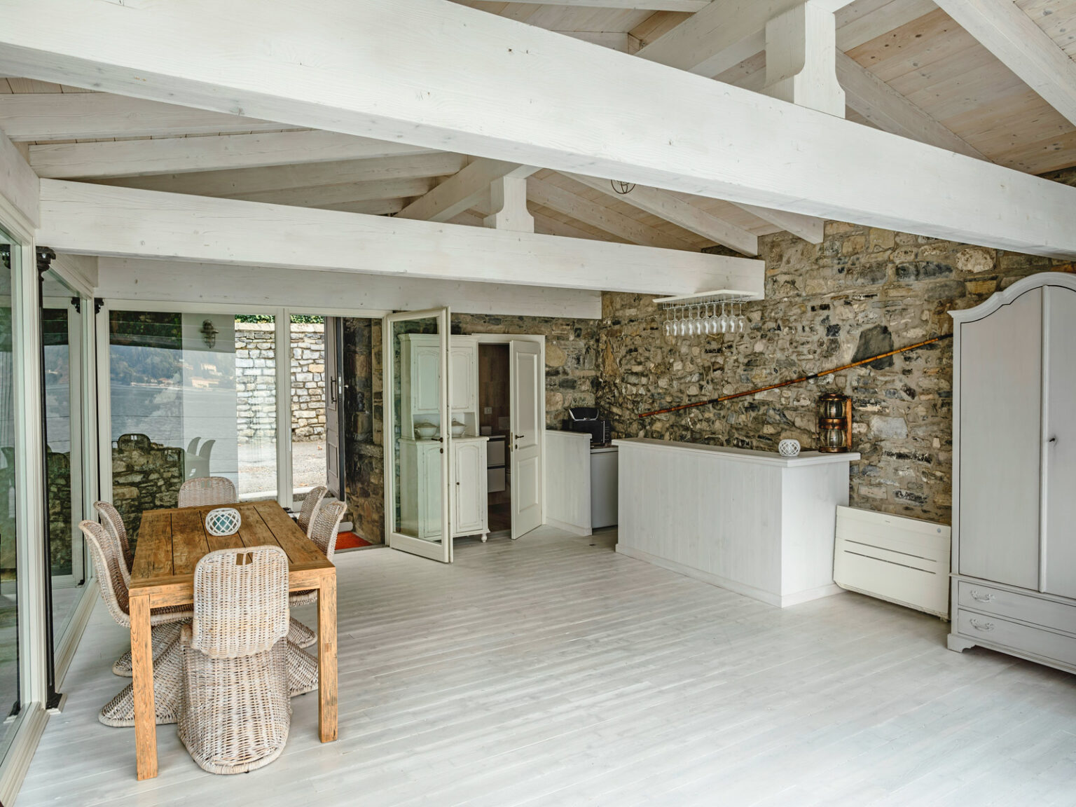 Bright and airy interior of a modern kitchen and dining area featuring wooden furniture, stone walls, and white ceiling beams. The space includes a dining table, a white kitchen counter, and large windows for natural light. | Only on SopranoVillas