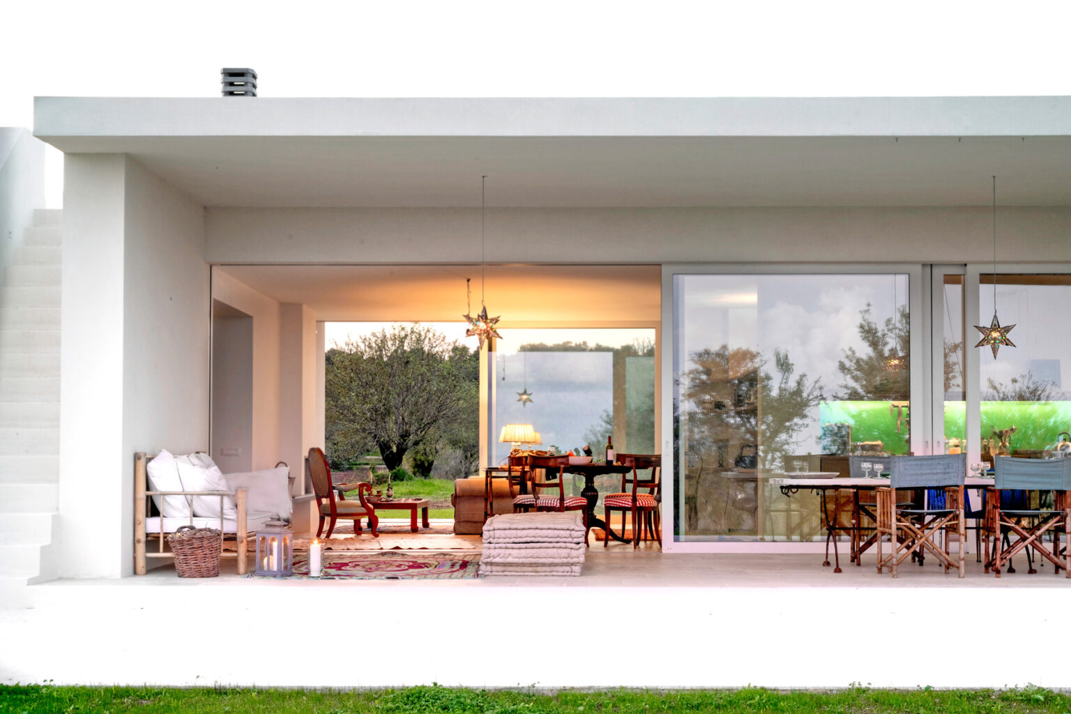 Modern open-concept home interior with large glass sliding doors, showcasing a stylish living area and dining space surrounded by greenery. Cozy seating arrangements and contemporary decor create a welcoming atmosphere. | Only on SopranoVillas