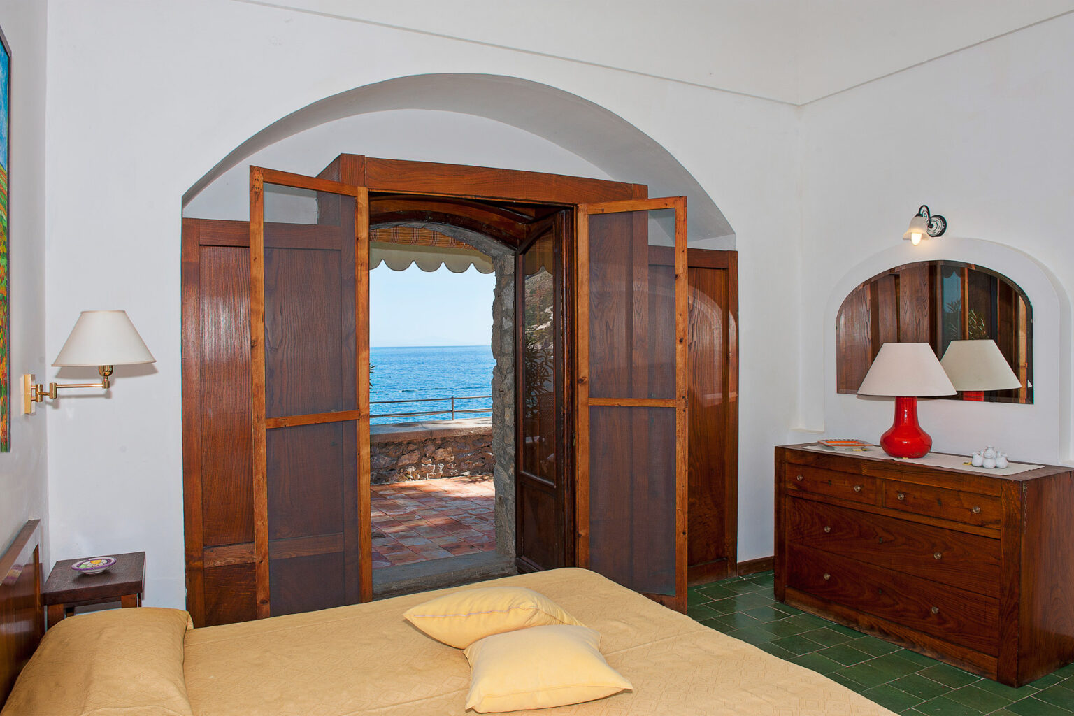 Cozy bedroom with a yellow bedspread, wooden furniture, and large open doors revealing a stunning ocean view. The room features a bedside lamp and a mirror, creating a warm and inviting atmosphere. | Only on SopranoVillas