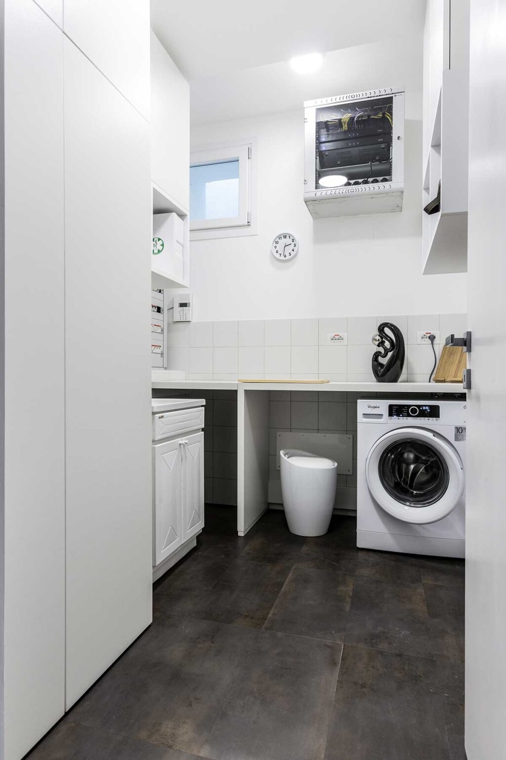 Modern laundry room featuring a washing machine, white cabinetry, a clock on the wall, and a trash bin, designed for efficiency and organization. | Only on SopranoVillas