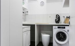 Modern laundry room featuring a washing machine, white cabinetry, a clock on the wall, and a trash bin, designed for efficiency and organization.