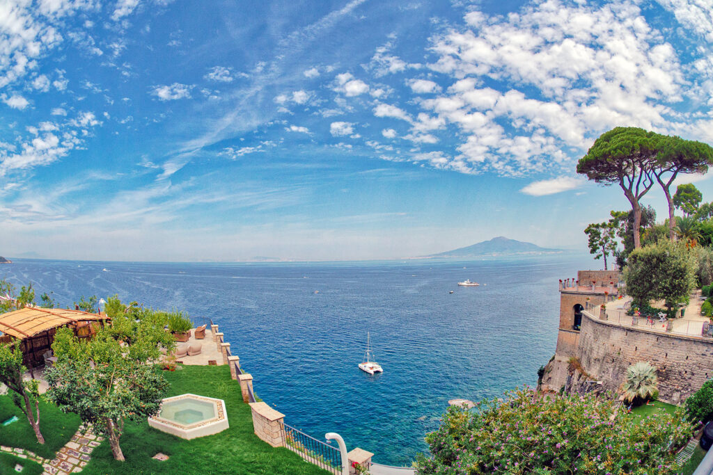 Scenic view of the coastline with lush greenery, a tranquil sea, and a distant mountain under a blue sky with clouds, showcasing a picturesque landscape ideal for travel and relaxation. | Only on SopranoVillas