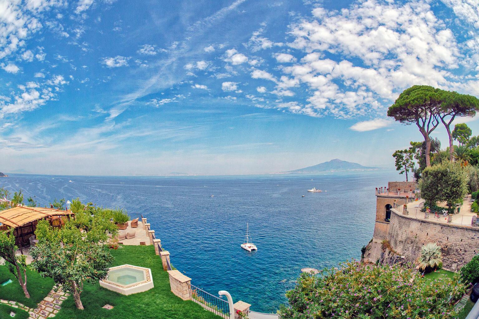 Scenic view of the coastline with lush greenery, a tranquil sea, and a distant mountain under a blue sky with clouds, showcasing a picturesque landscape ideal for travel and relaxation. | Only on SopranoVillas