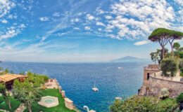 Scenic view of the coastline with lush greenery, a tranquil sea, and a distant mountain under a blue sky with clouds, showcasing a picturesque landscape ideal for travel and relaxation.