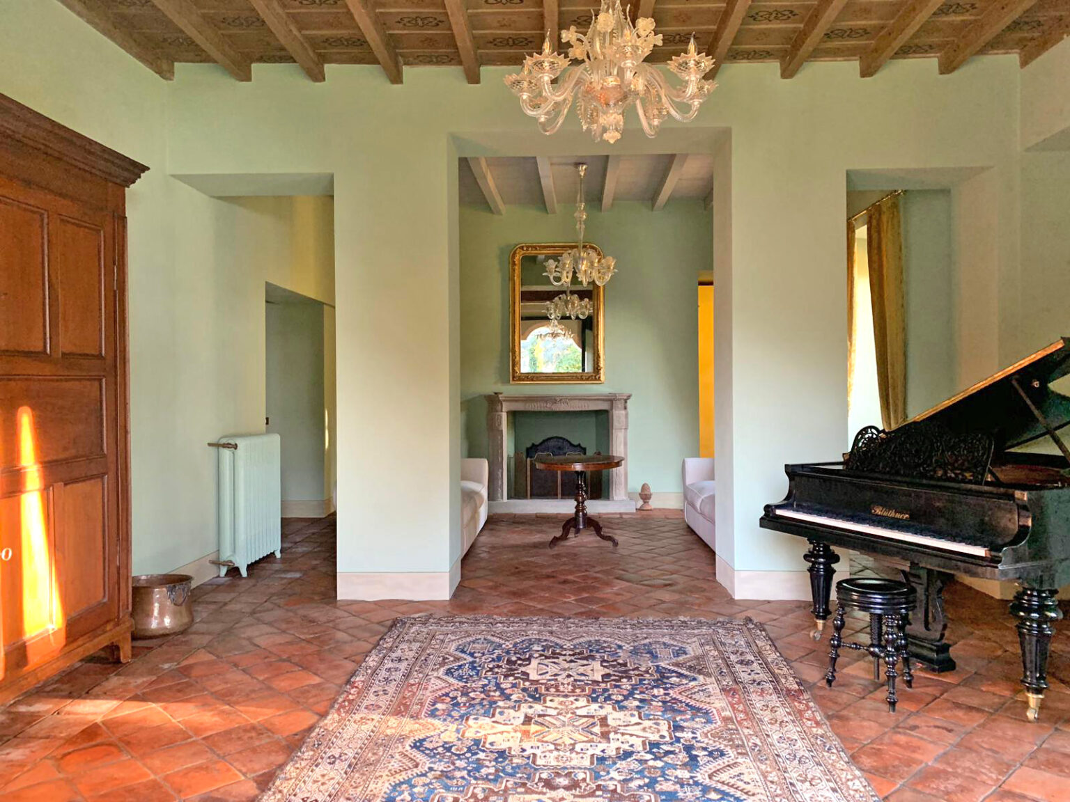 Elegant living room featuring a grand piano, vintage chandelier, and classic decor. The space is illuminated by natural light, highlighting the charming details such as a fireplace and decorative mirror. | Only on SopranoVillas