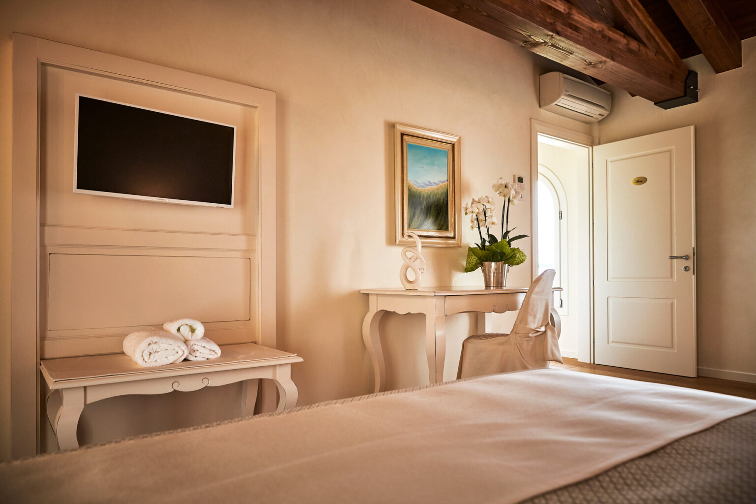 Cozy hotel room featuring a queen-size bed, a flat-screen TV mounted on the wall, a white desk with a decorative plant, and a door leading outside. The warm, inviting decor includes wooden beams and soft lighting, creating a relaxing atmosphere. | Only on SopranoVillas