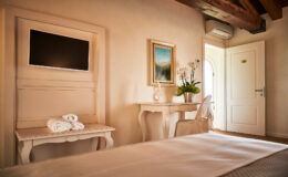 Cozy hotel room featuring a queen-size bed, a flat-screen TV mounted on the wall, a white desk with a decorative plant, and a door leading outside. The warm, inviting decor includes wooden beams and soft lighting, creating a relaxing atmosphere.