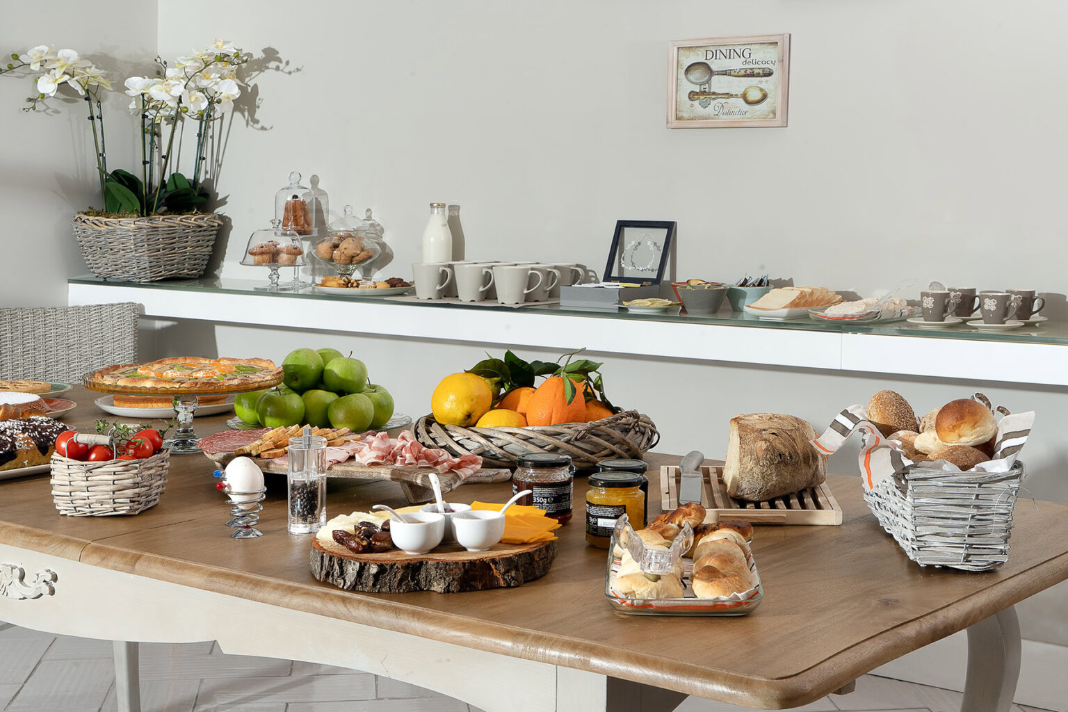 A beautifully arranged breakfast spread featuring a variety of fresh fruits, pastries, bread, and cold cuts on a wooden table, with decorative baskets and a minimalist background. | Only on SopranoVillas