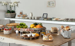 A beautifully arranged breakfast spread featuring a variety of fresh fruits, pastries, bread, and cold cuts on a wooden table, with decorative baskets and a minimalist background.