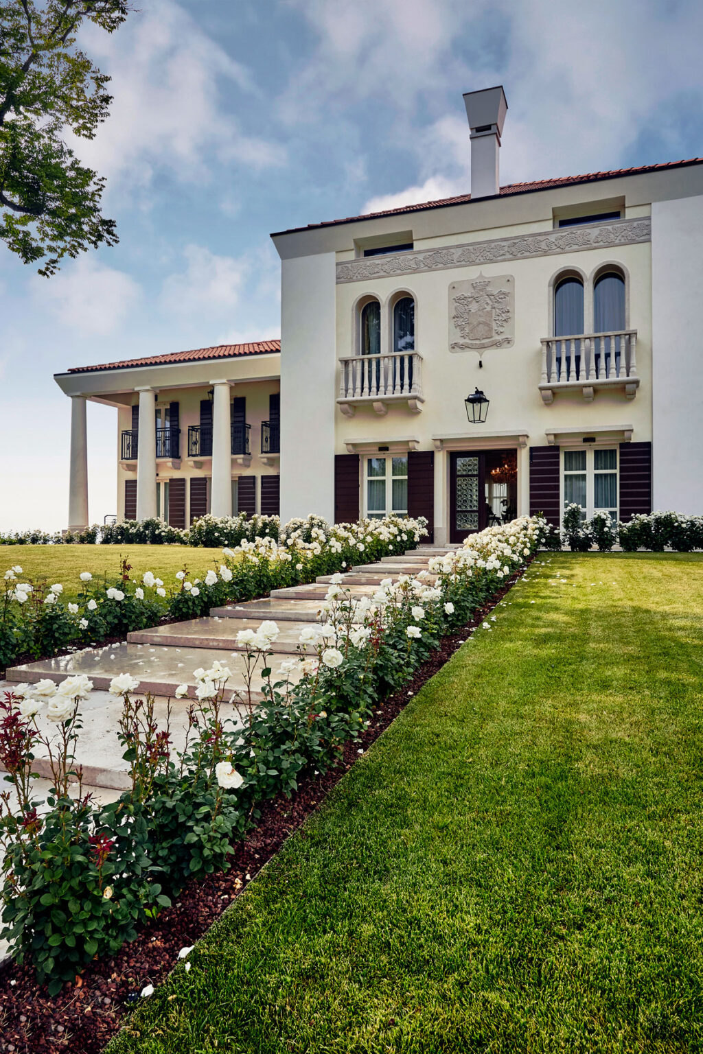 Elegant modern mansion with a landscaped garden featuring a pathway lined with white roses, showcasing classic architectural elements and a welcoming entrance. | Only on SopranoVillas