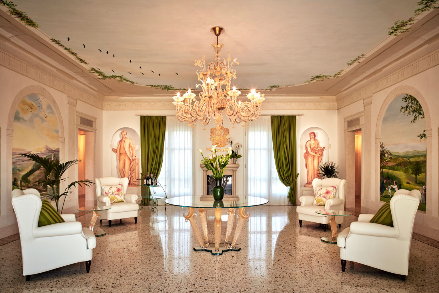 Elegant living room featuring a grand chandelier, two white armchairs, and a glass table adorned with flowers. The walls are decorated with soft pastel murals and lush green curtains, creating a luxurious and inviting atmosphere. | Only on SopranoVillas