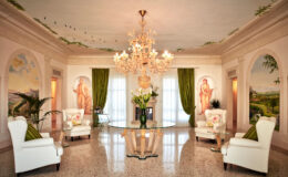 Elegant living room featuring a grand chandelier, two white armchairs, and a glass table adorned with flowers. The walls are decorated with soft pastel murals and lush green curtains, creating a luxurious and inviting atmosphere.