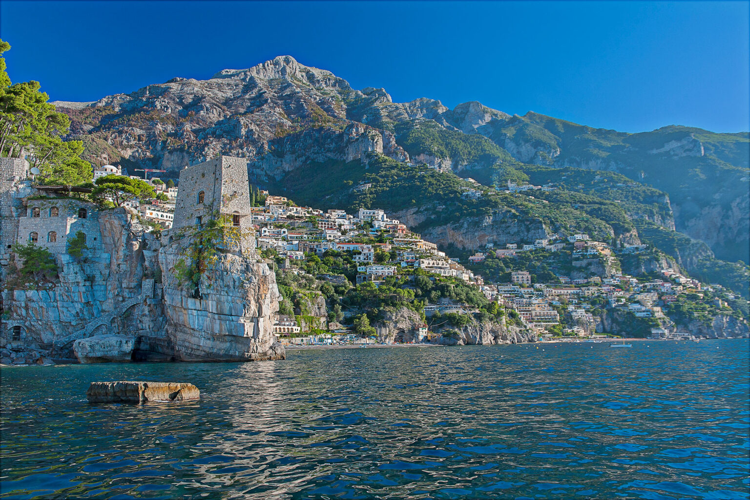 Scenic view of the Amalfi Coast with rocky cliffs, historic tower, and colorful buildings nestled in the hillside under a clear blue sky. | Only on SopranoVillas