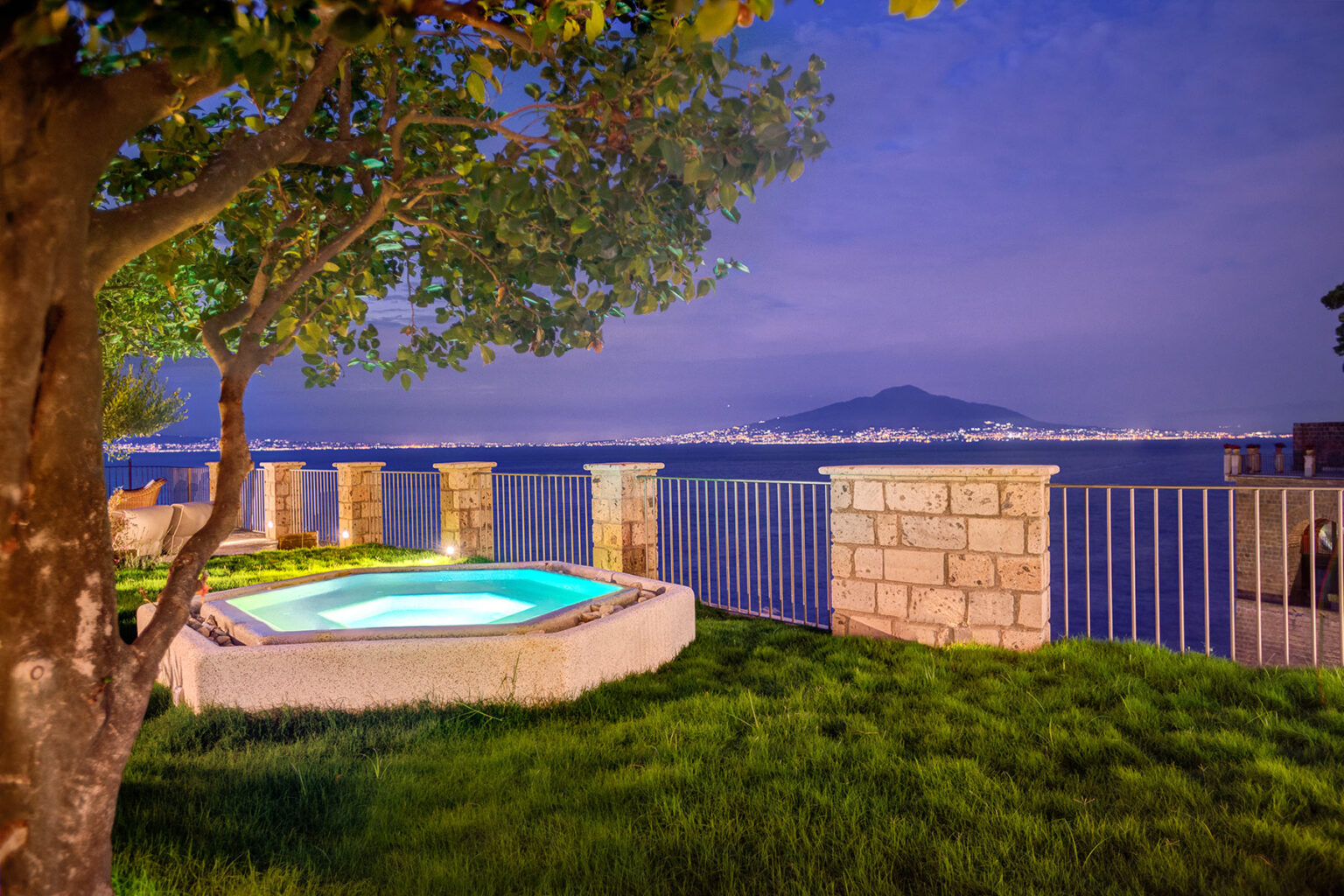 Scenic view of a hot tub beside a lush green lawn, overlooking a serene waterfront at dusk, with distant hills illuminated by city lights in the background. | Only on SopranoVillas