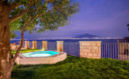 Scenic view of a hot tub beside a lush green lawn, overlooking a serene waterfront at dusk, with distant hills illuminated by city lights in the background.