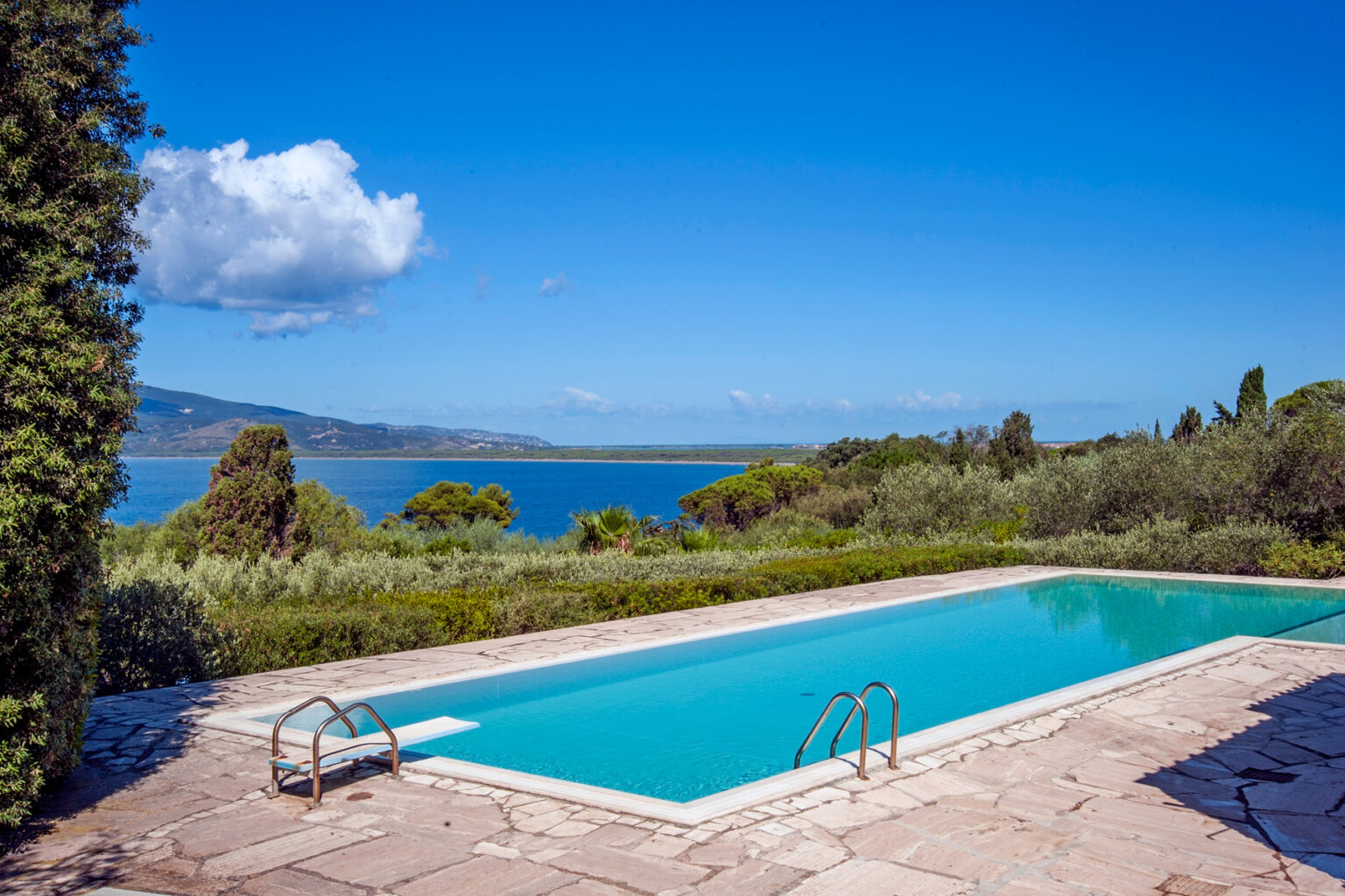 Scenic view of a serene swimming pool surrounded by lush greenery, with a clear blue sky and distant ocean in the background. Ideal for vacation and relaxation. | Only on SopranoVillas