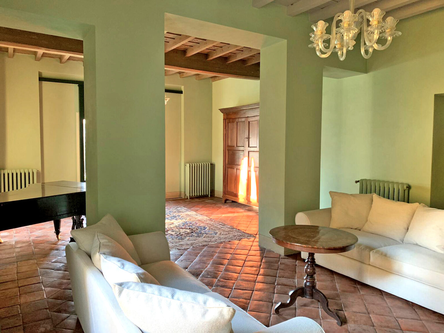 Spacious living room featuring light green walls, a cozy white sofa, and a wooden coffee table, with natural light streaming in through large windows, showcasing rustic tile flooring and elegant chandelier. | Only on SopranoVillas