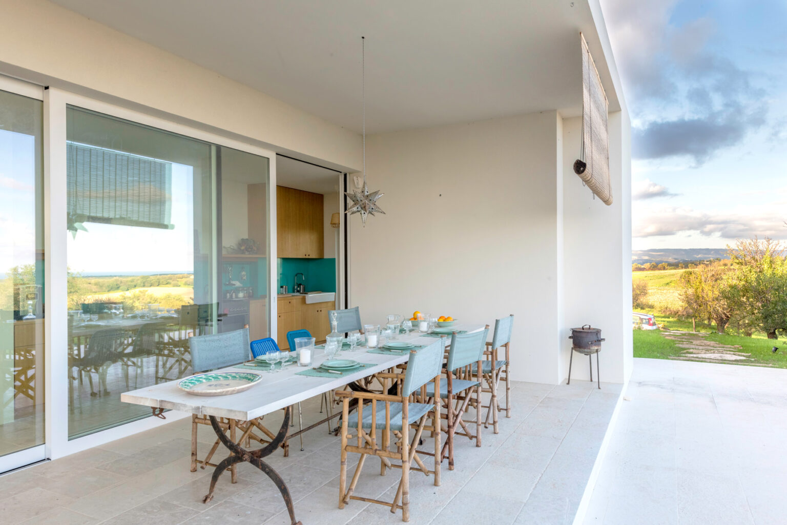 Modern outdoor patio dining area with a large table set for meals, featuring blue and white tableware, surrounded by a serene landscape view. Large glass doors lead to an indoor space, enhancing the open-air feel. | Only on SopranoVillas